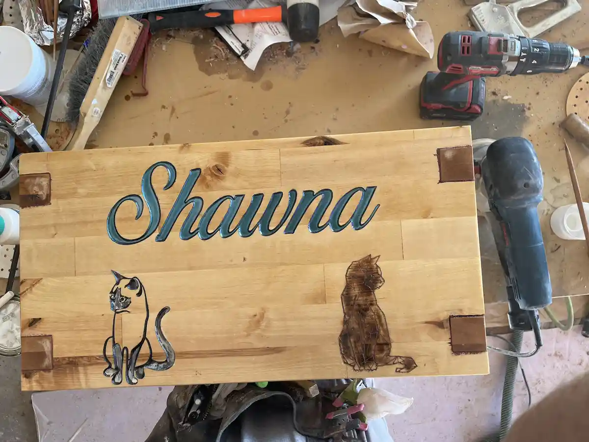 A wooden sign with the name 'Shawna' in teal cursive lettering, flanked by two stylized cat silhouettes—one metallic and one…