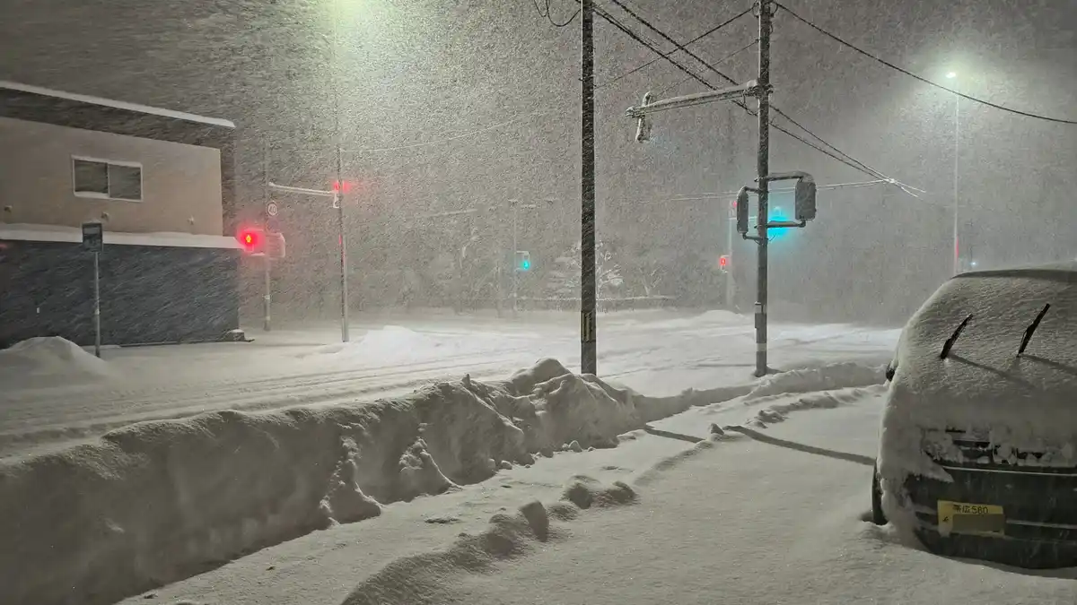 A nighttime street scene during a heavy snowstorm, with snow-covered cars, traffic lights glowing red and green, and…