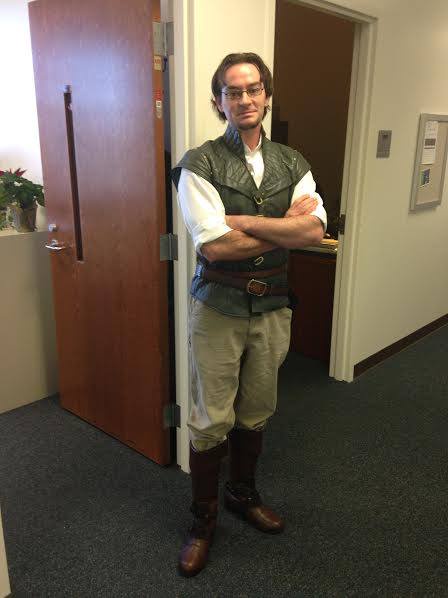 A man in a modern office hallway, dressed in a fantasy adventurer costume with a vest, belt, boots, and glasses, stands with…