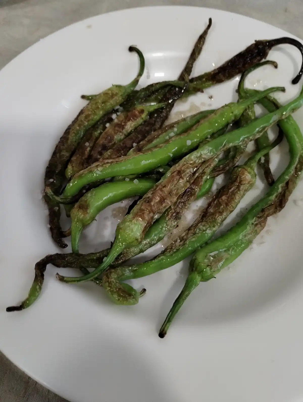 A white plate holds a pile of roasted green chili peppers, some charred and blistered, sprinkled with coarse salt. The…