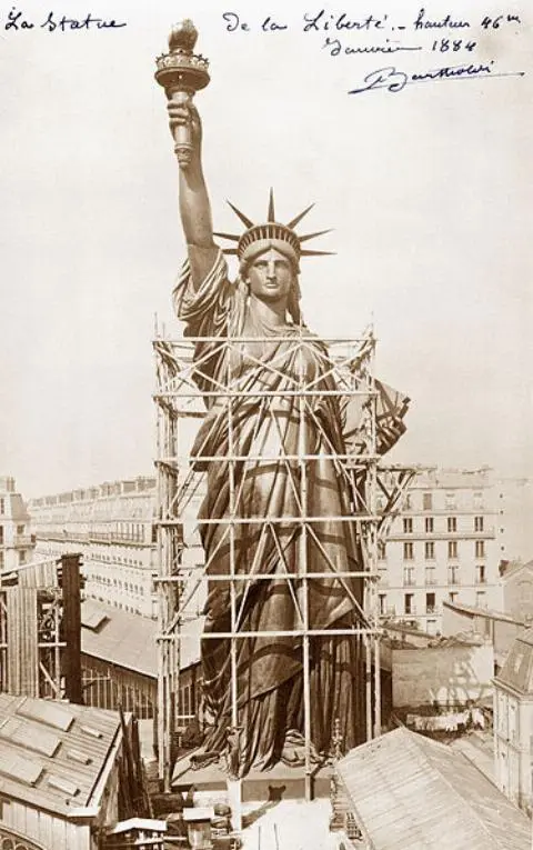 A sepia-toned historical photograph shows the Statue of Liberty under construction, surrounded by scaffolding, with…