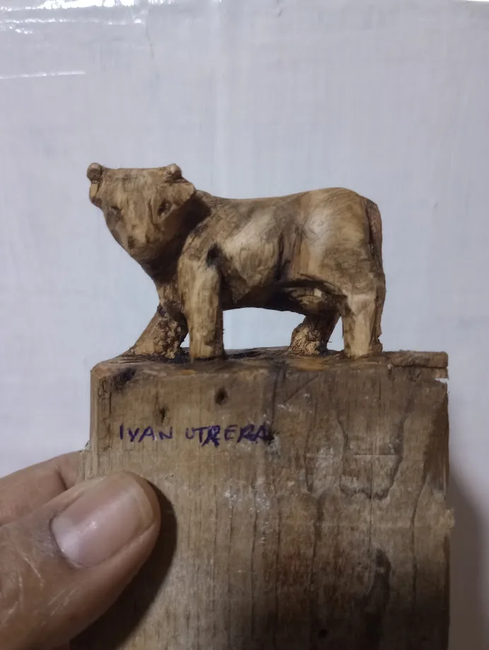 A hand holds a rough wooden carving of a bull standing on a thick wooden block. The name 'IYAN UTRERA' is handwritten in…