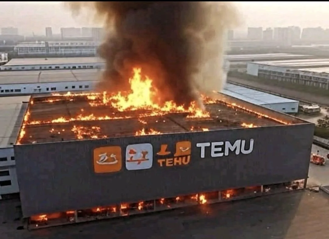 An aerial view shows a large industrial building engulfed in flames and thick black smoke, with the side of the building…