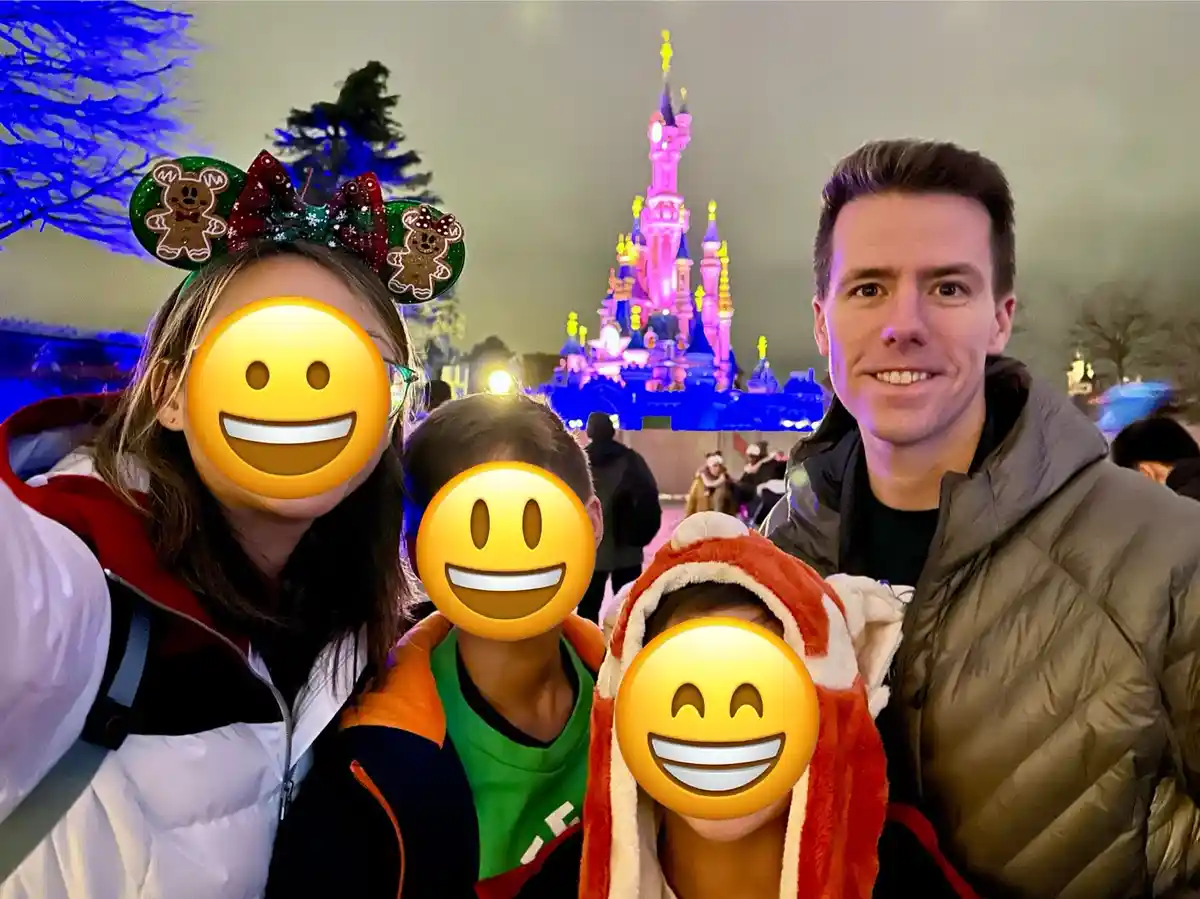 A family selfie at a Disney park at night, with the castle lit up in the background. Three children have their faces…
