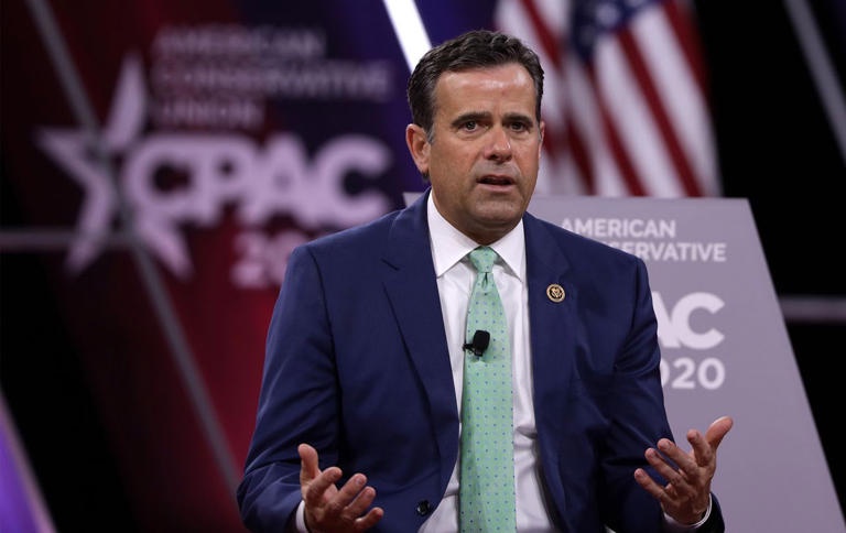 a man in a blue suit and light green tie stands at a CPAC 2020 event, gesturing with both hands while speaking. Behind him…