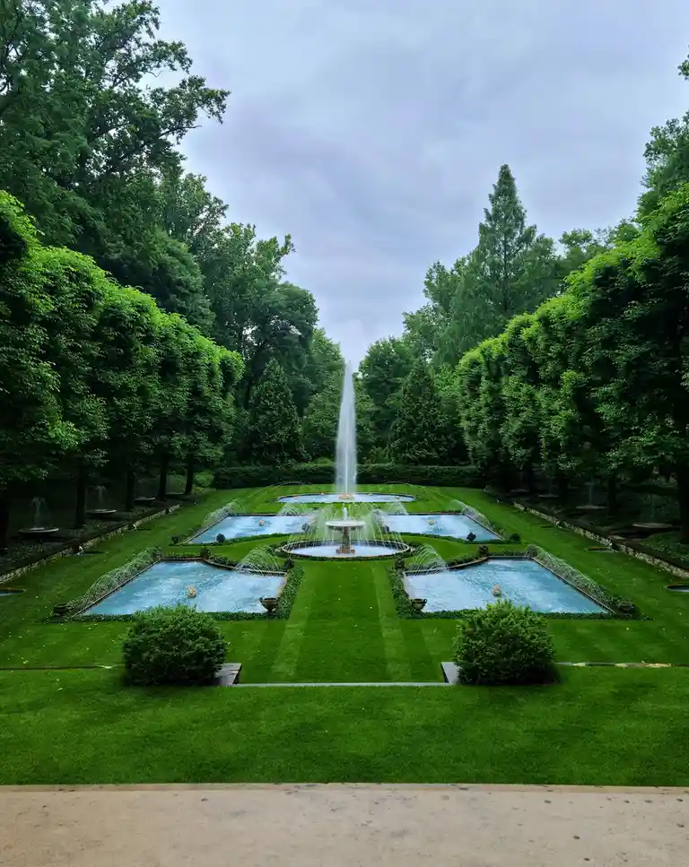 A symmetrical, ornate garden with a central fountain shooting water high into the air, surrounded by manicured lawns…