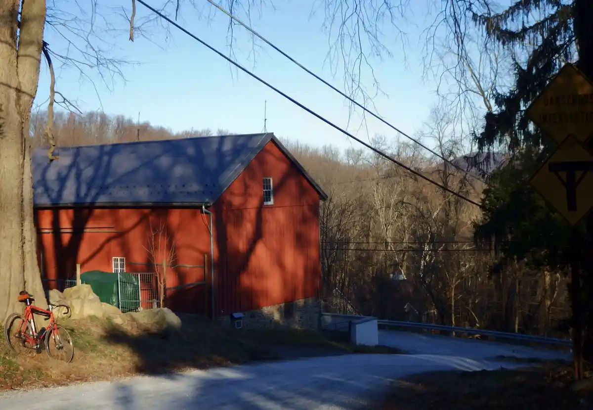 A red barn with a dark roof stands beside a winding road under a clear blue sky, with bare trees casting long shadows. A…