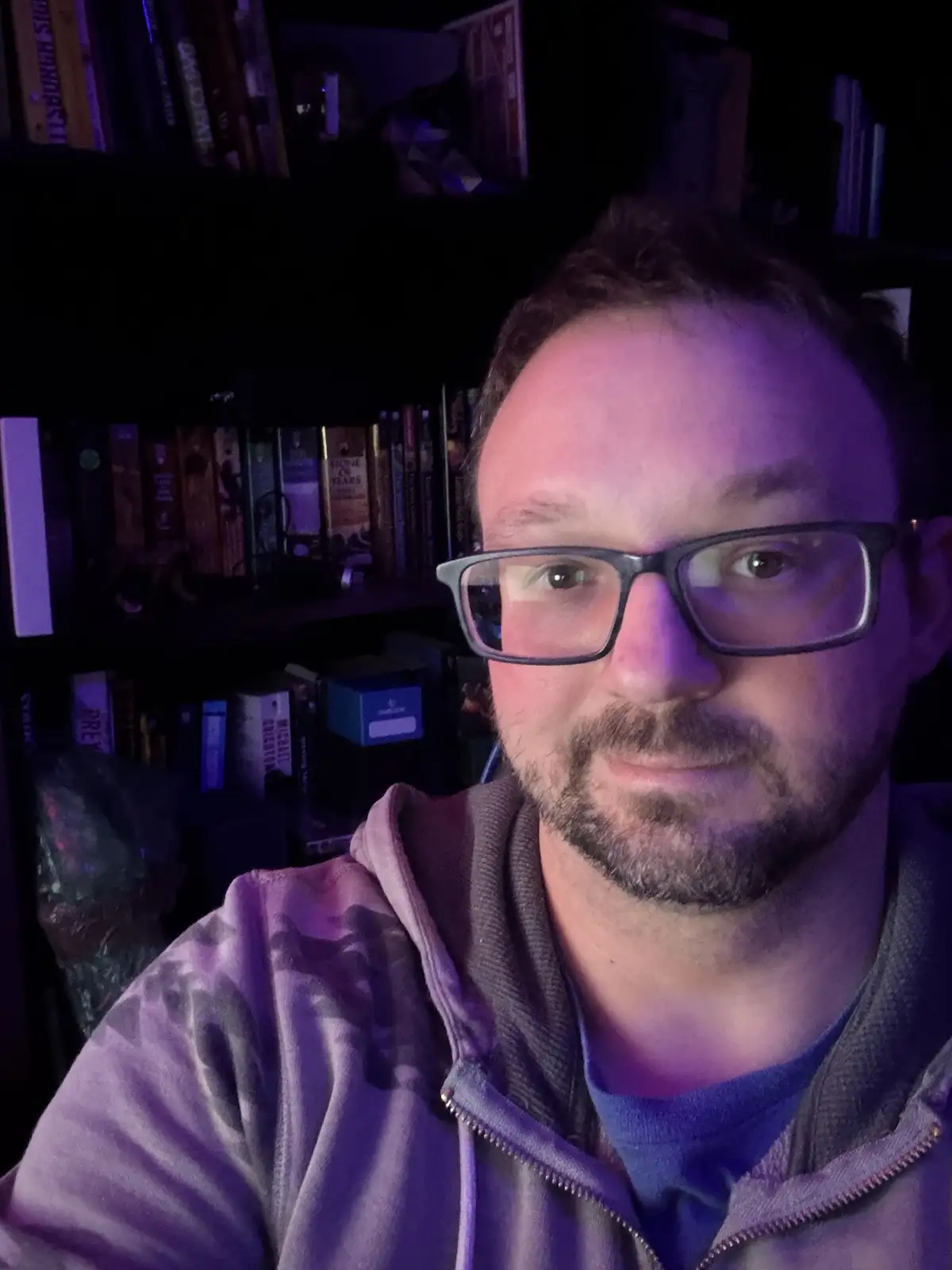 A man with glasses and a beard, wearing a zip-up hoodie, looks directly at the camera with a slightly smug or unimpressed…