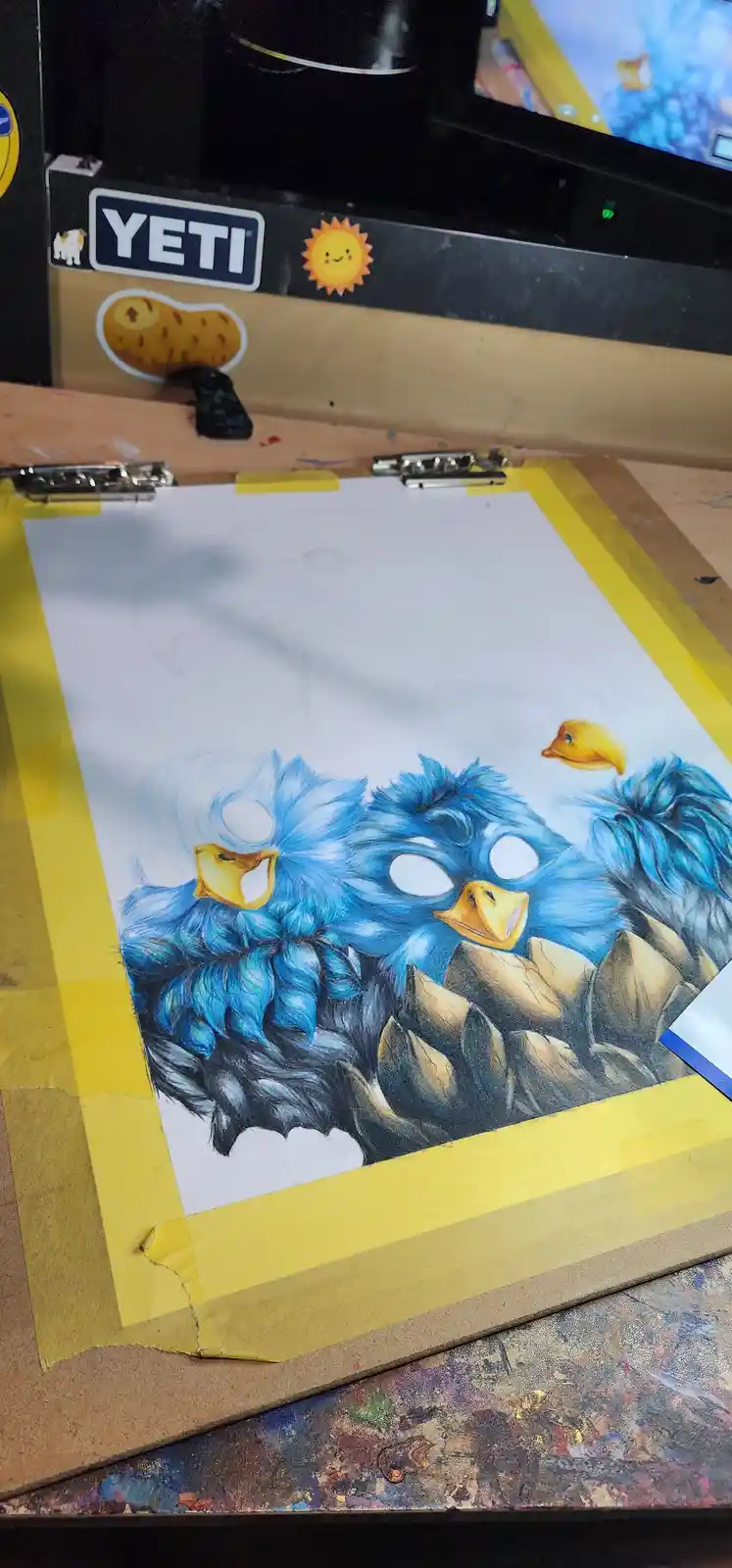 A partially completed colored pencil drawing of three fluffy blue birds with yellow beaks, perched on rocks, is taped to a…