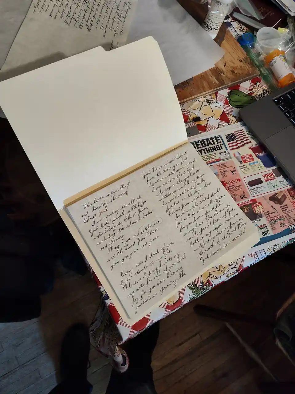 An overhead shot shows a handwritten letter in a folder, placed on a cluttered table with a newspaper, laptop, and various…