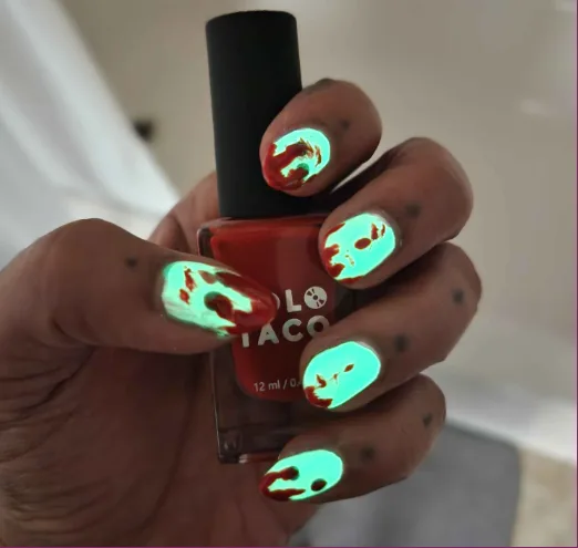 A hand holds a red nail polish bottle labeled 'POLO TACO' while showcasing nails painted with glowing green and red designs…