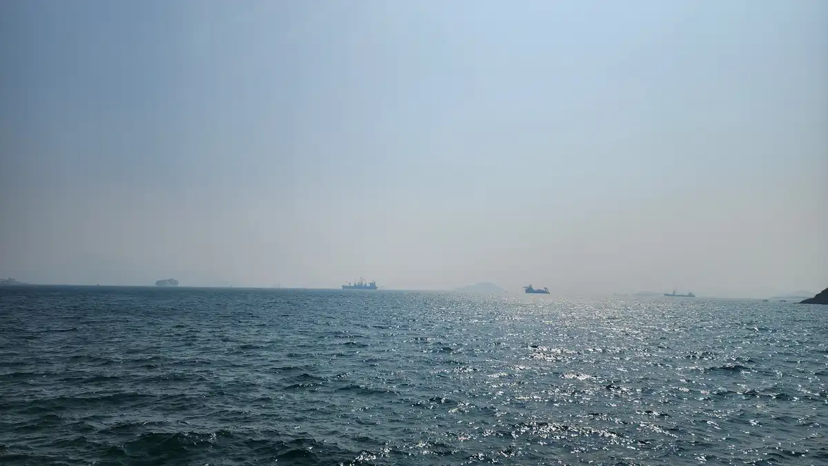A wide shot of a calm, sunlit ocean with several large cargo ships visible on the horizon under a hazy sky. The overlaid…