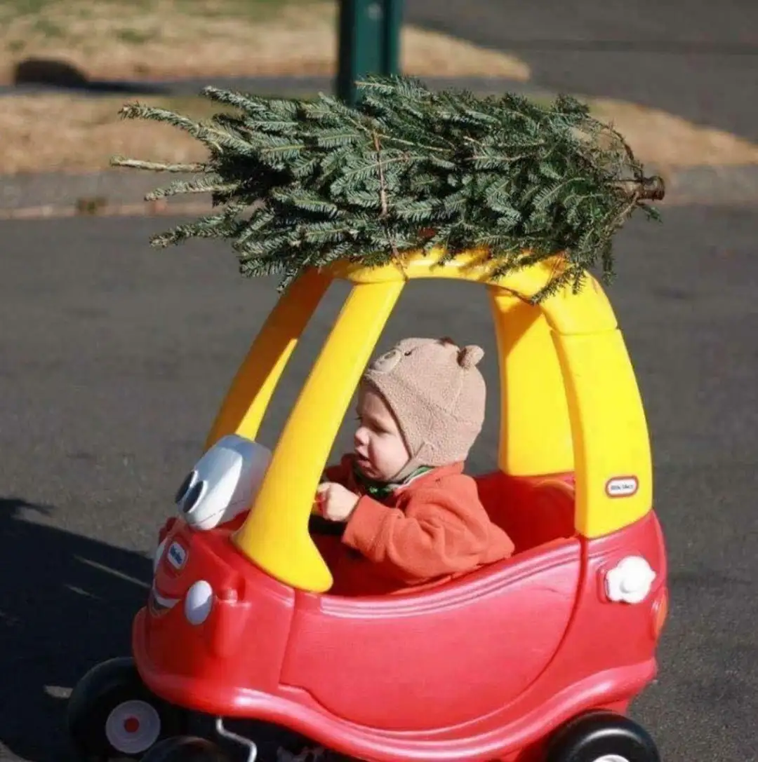 A toddler wearing a brown bear hat and red jacket sits in a red and yellow Little Tikes Cozy Coupe toy car, looking forward…
