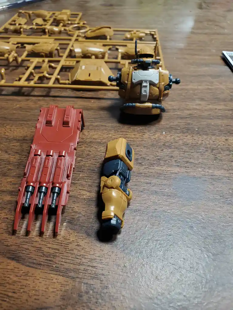 A photo shows unassembled plastic robot parts on a wooden table, including a red claw-like hand, a tan and black robotic…