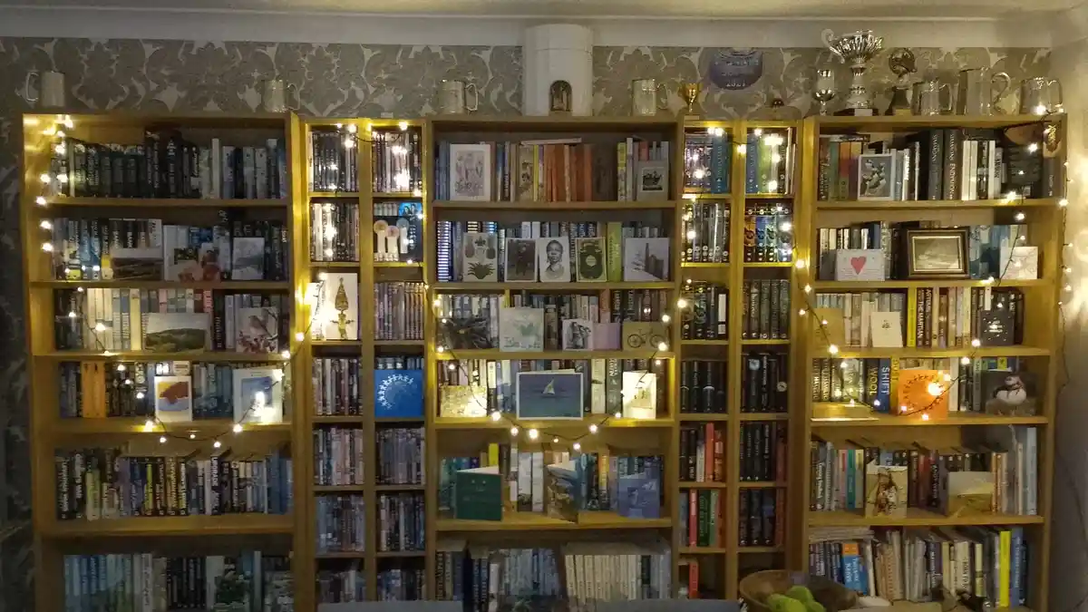 A wide shot of a large, wooden bookshelf filled with numerous books, decorated with string lights and various small items…