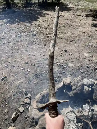 A hand holds up a long, gnarled stick that resembles a sword, standing upright over a smoldering campfire pit with rocks and…