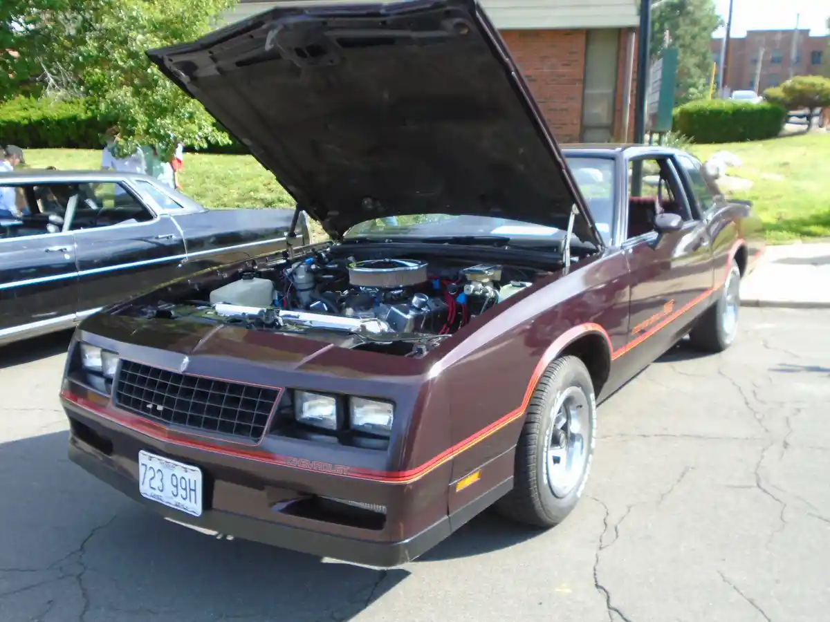 A maroon Chevrolet Monte Carlo SS with its hood open, revealing a modified engine, parked on cracked asphalt with another…