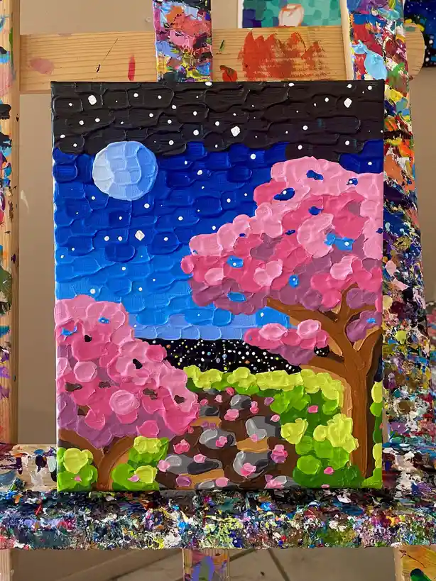 A thickly textured, colorful painting on a canvas depicts two pink cherry blossom trees under a blue and black night sky…