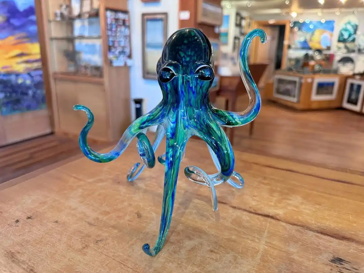 A glass sculpture of an octopus with swirling blue and green patterns stands on a wooden table in what appears to be an art…