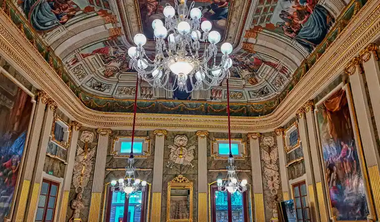 An ornate, opulent room with a grand chandelier, gilded moldings, marble walls, and classical frescoes on the ceiling. The…
