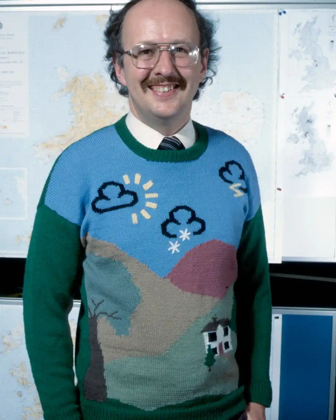 A smiling man with glasses and a mustache wears a colorful, knitted sweater depicting a weather scene with sun, clouds…