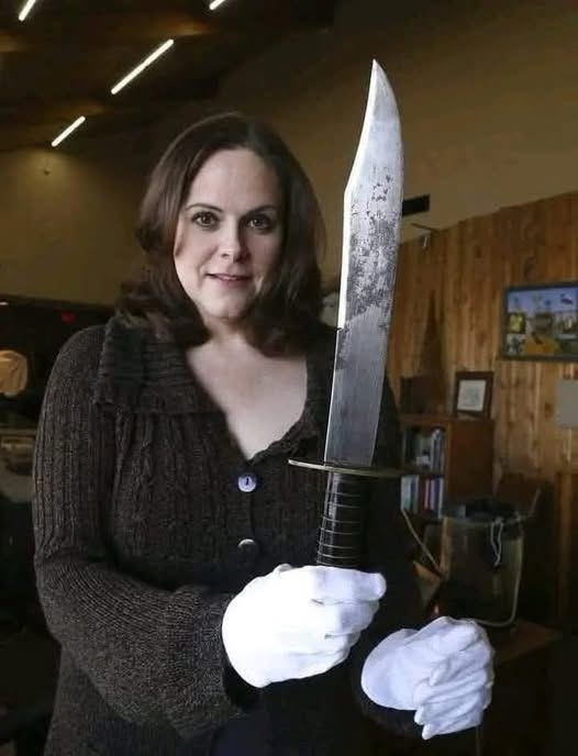 a woman with shoulder-length brown hair wearing a dark knit cardigan and white gloves holds a large, dirty-looking knife…