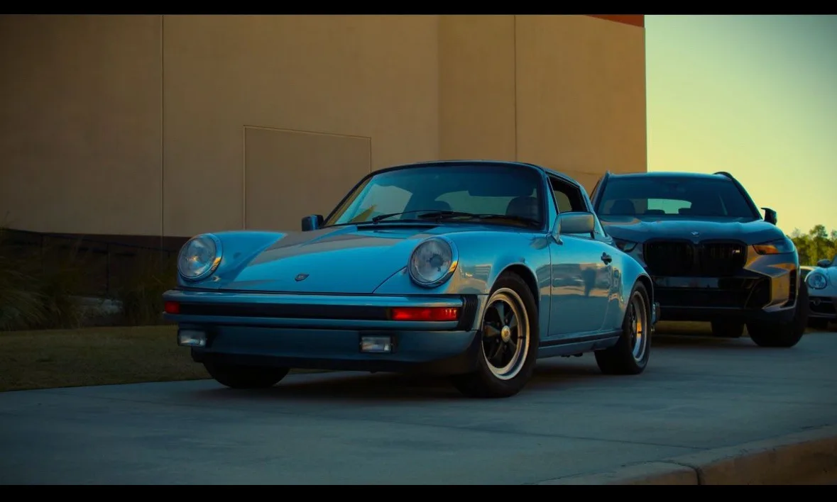 A light blue classic Porsche 911 is parked prominently in the foreground, with a modern black BMW SUV visible behind it. The…