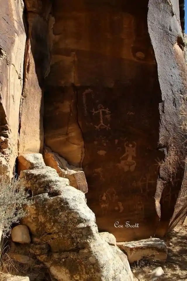 A photograph of ancient rock art, or petroglyphs, carved into a dark brown sandstone cliff face within a narrow canyon. The…