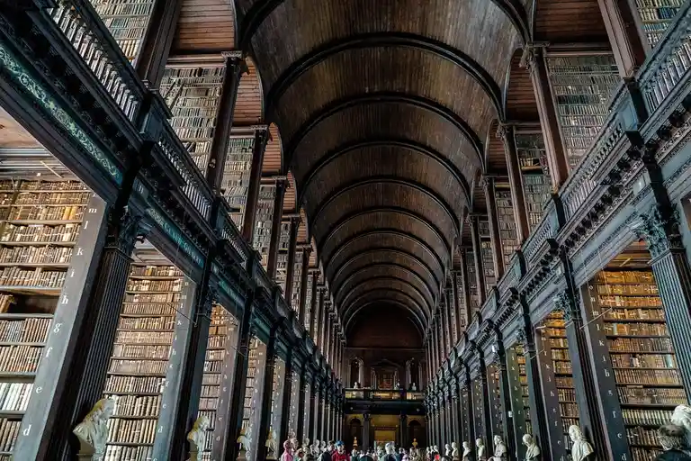 A grand, ornate library with towering wooden bookshelves lining both sides of a long, arched hallway, filled with visitors…
