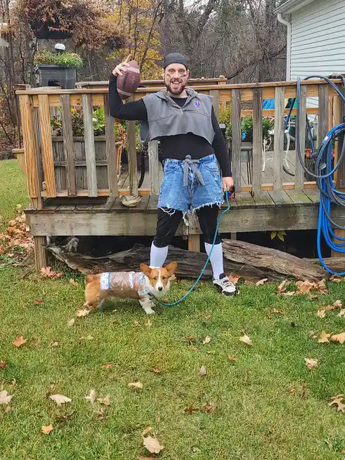 A man dressed in football gear, including face paint, a vest, and shorts, stands on a wooden deck holding a football while…