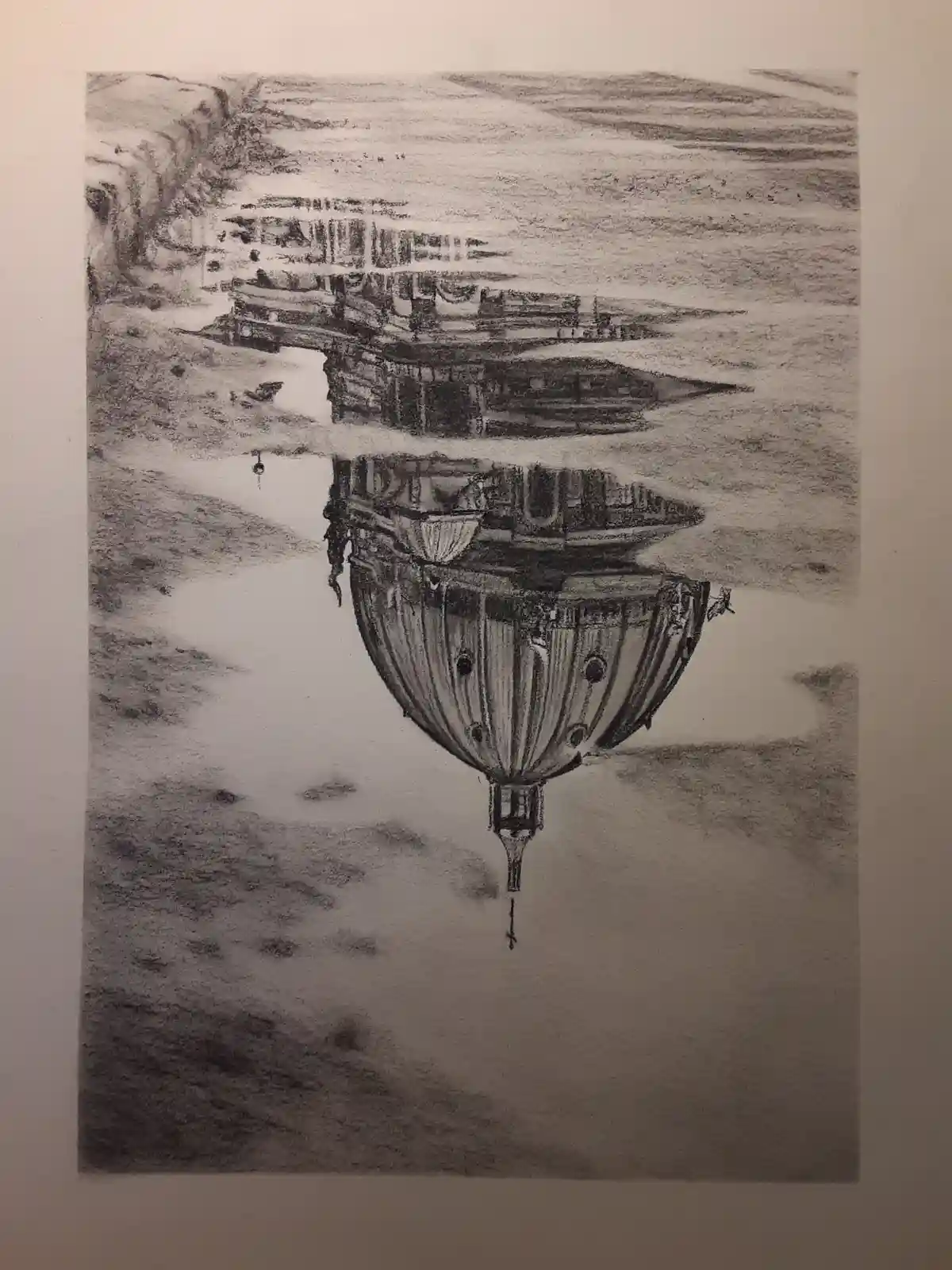 A black-and-white pencil drawing shows a ship on water with its reflection distorted into a hot air balloon shape below the…