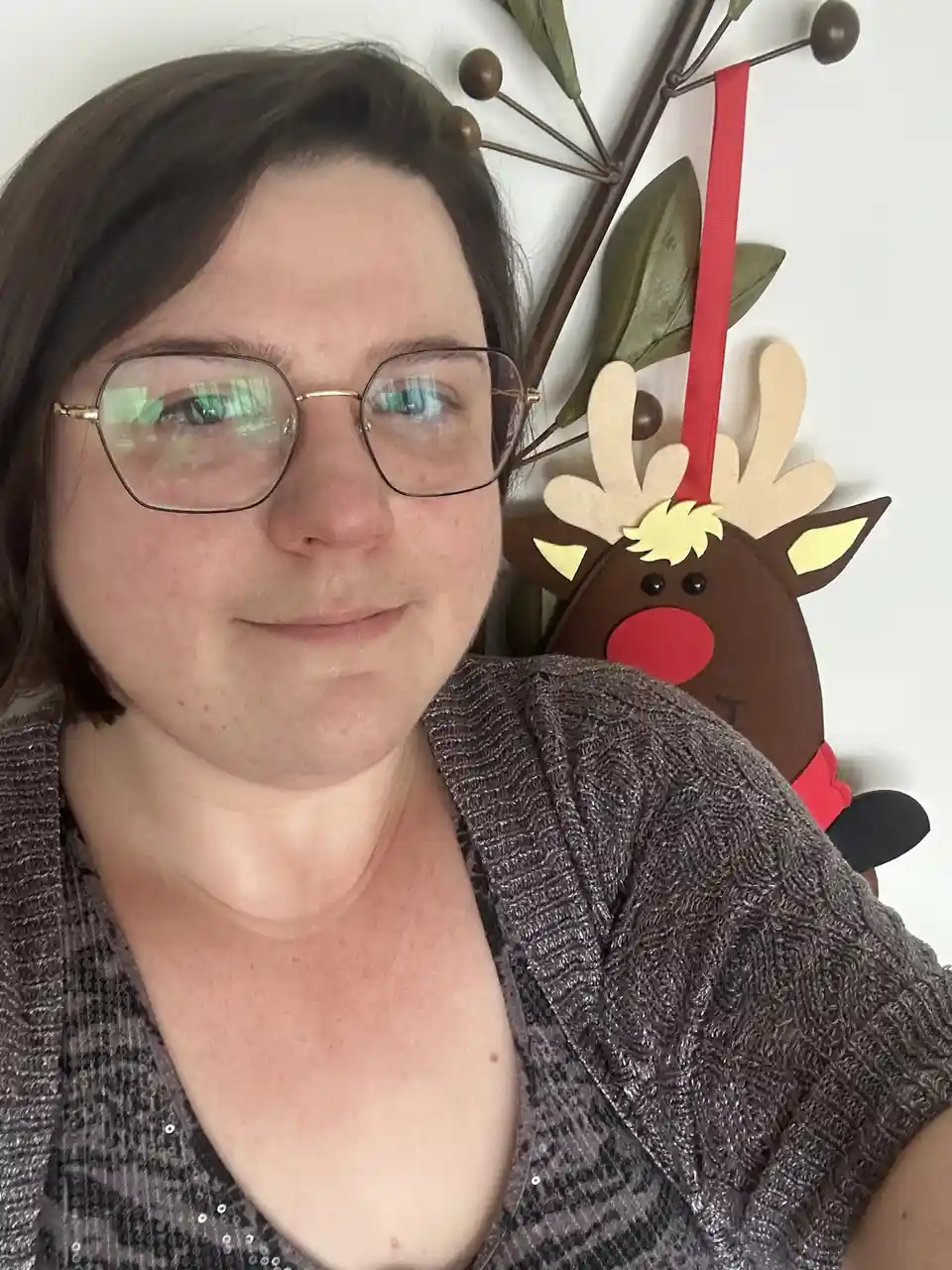 A person with glasses and a sparkly gray top smiles at the camera, standing in front of a wall decoration featuring a brown…