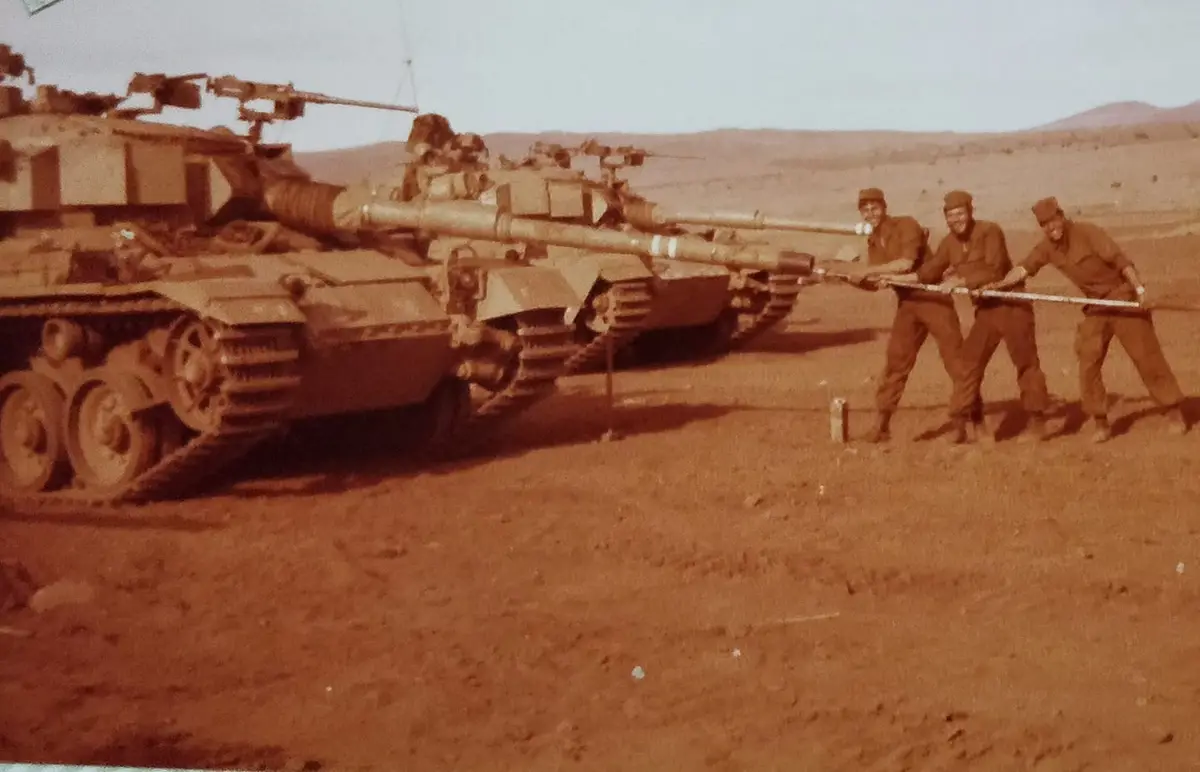 Three smiling soldiers in military uniforms are pulling on a long pole attached to the barrel of a large tank in a desert…