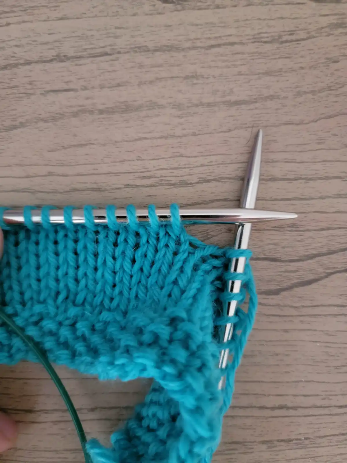 A close-up photo shows a turquoise knitted fabric on two silver knitting needles, resting on a light wood-grain surface. The…