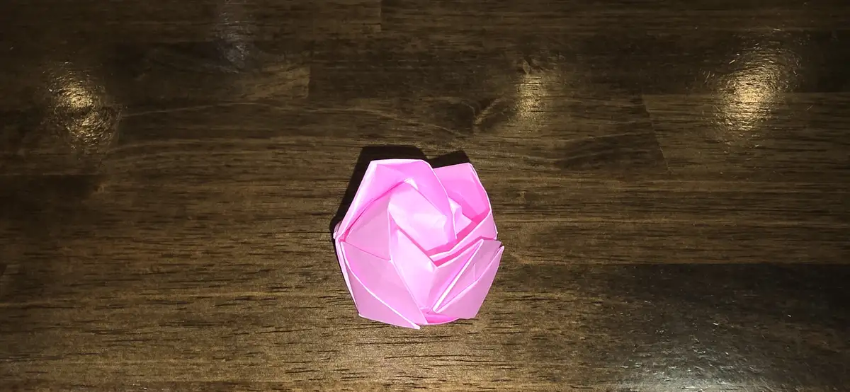 A pink origami rose sits centered on a dark wooden surface, its intricate folds catching the light. The overlaid text reads…
