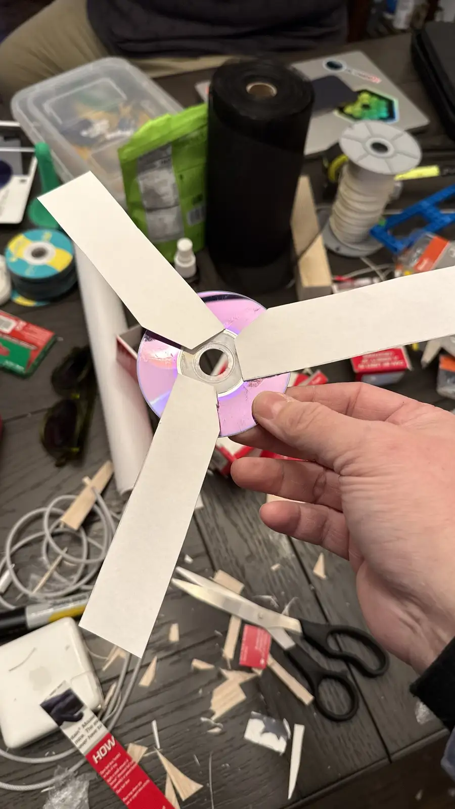A hand holds a makeshift fan prop made from a CD and three paper blades, surrounded by crafting supplies like scissors, wood…