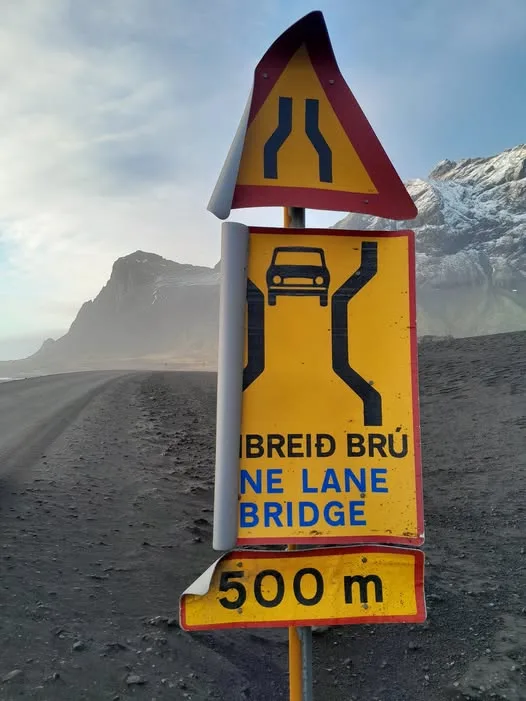 A roadside sign in Iceland warns of an upcoming one-lane bridge 500 meters ahead, featuring a triangular warning symbol…