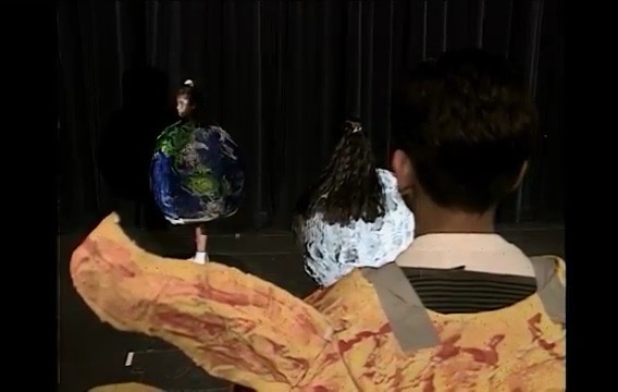 A person in a large, colorful costume resembling a planet stands on a stage, viewed from behind by another person in a…