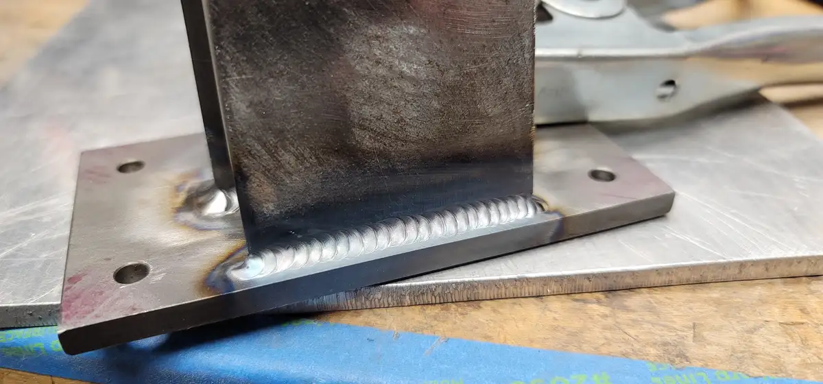 A close-up photo shows a metal weld joining a vertical steel bar to a horizontal steel plate, with visible weld beads and…
