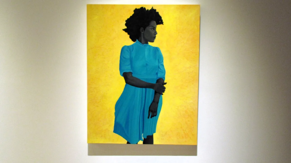 A large painting hangs on a white wall, depicting a Black woman in profile wearing a bright blue dress against a textured…