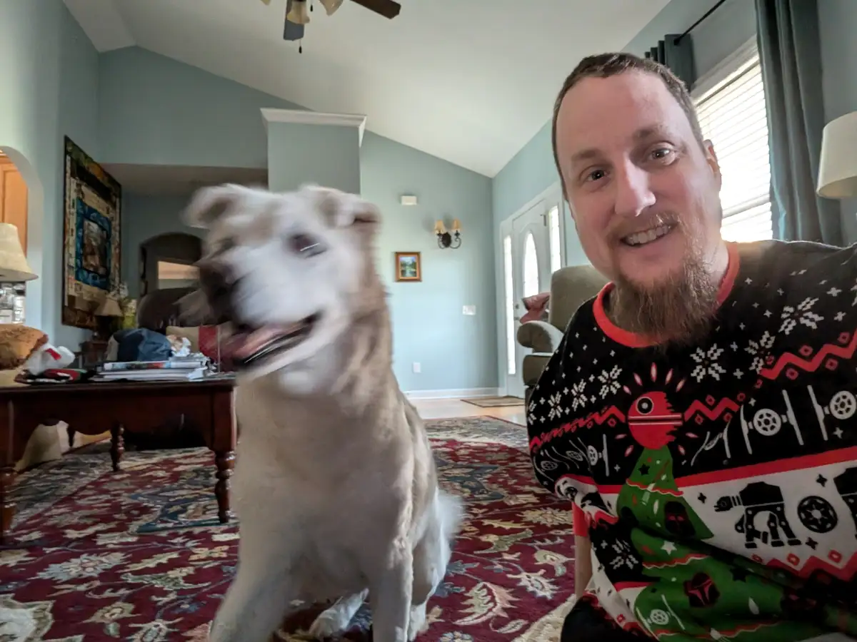 A man with a beard wearing a Star Wars-themed Christmas sweater smiles at the camera while a white dog with motion blur…