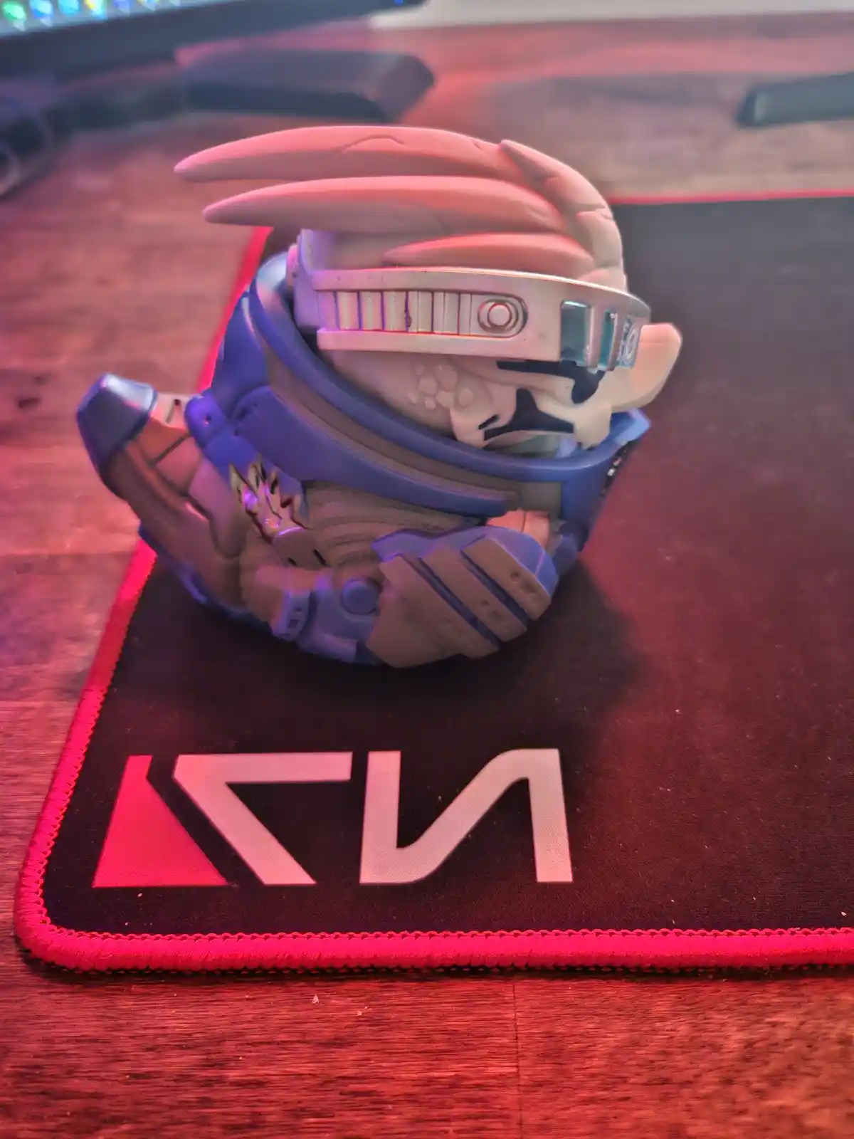 A figurine of a character with a helmet and blue armor sits on a black mousepad with the 'KIA' logo, under red ambient…
