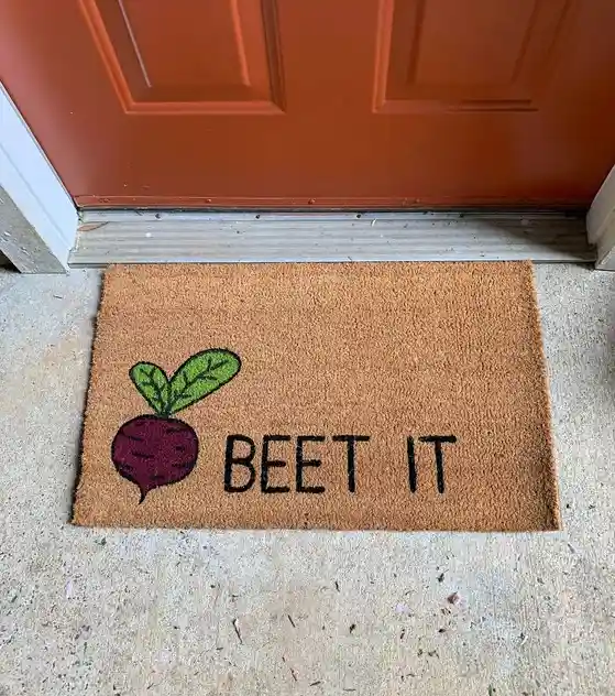 A brown doormat with a cartoon beetroot and the text 'BEET IT' sits on a concrete porch in front of a reddish-brown door…