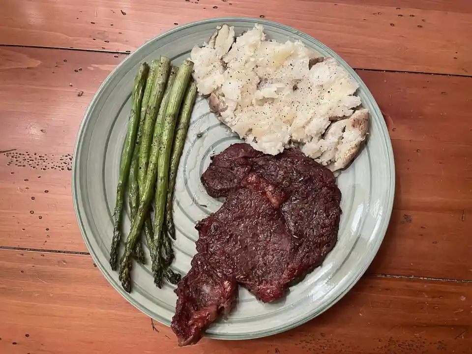 A plate of food on a wooden table contains a large, darkly seared steak, a serving of mashed potatoes with black pepper, and…