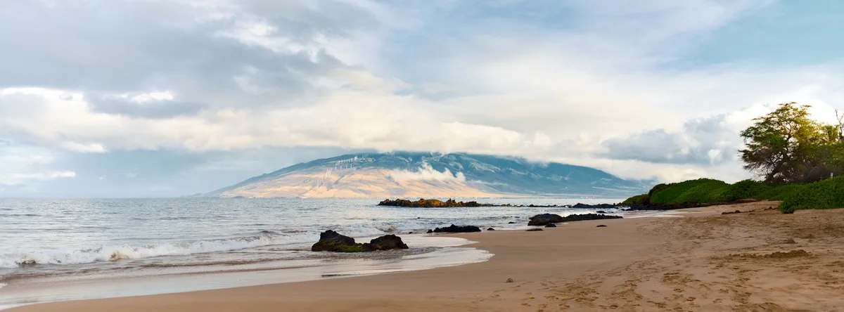 A serene beach panorama with gentle waves, sandy shore, and a distant mountain range partially obscured by clouds. On the…