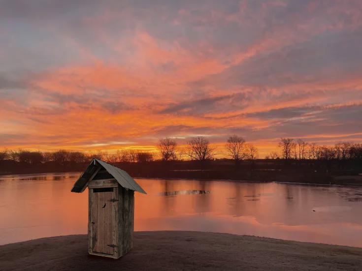 a rustic wooden outhouse stands alone on a lakeshore under a vibrant, fiery sunset sky with orange, pink, and purple clouds…