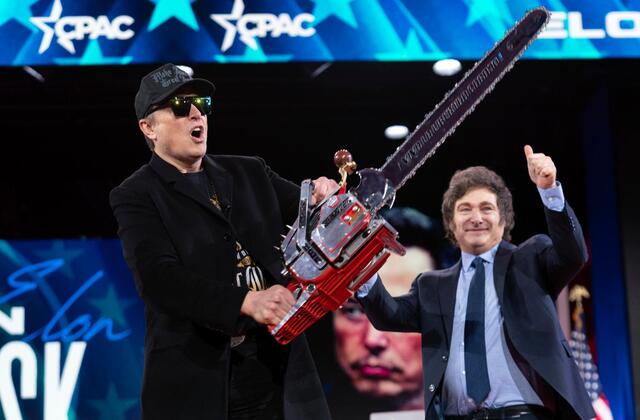 A man in a black suit and cap holds a large red chainsaw while shouting, standing next to another man in a suit who is…