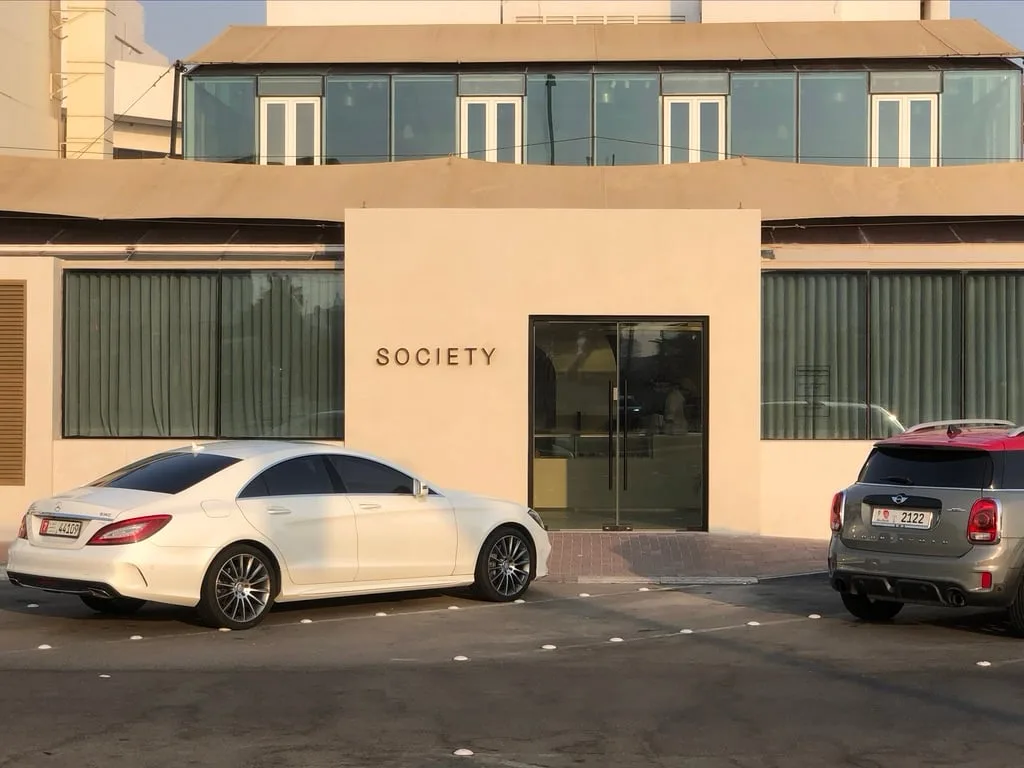 A beige building with a glass door and windows, labeled 'SOCIETY' on its facade, has two cars parked in front: a white…