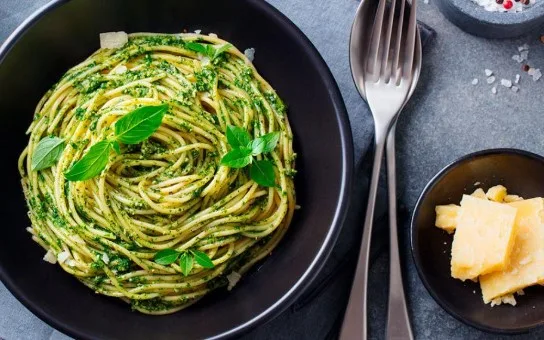 a close-up photo of a black bowl filled with spaghetti coated in green pesto sauce and garnished with fresh basil leaves. To…