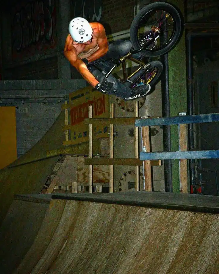 A shirtless man wearing a white helmet and black gloves is mid-air on a BMX bike, performing a trick above a wooden ramp…